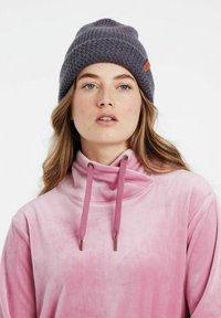 Beanie grigia lavorata a maglia con un motivo strutturato e un accento in pelle. Il modello indossa una felpa rosa in velluto con dettagli a coulisse.