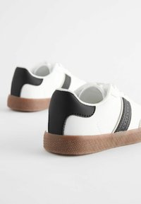 Białe sneakersy z czarnymi skórzanymi akcentami, strukturalnymi brązowymi gumowymi podeszwami i dwoma czarnymi paskami po bokach. Okrągły czubek i wyściełane wnętrze.
