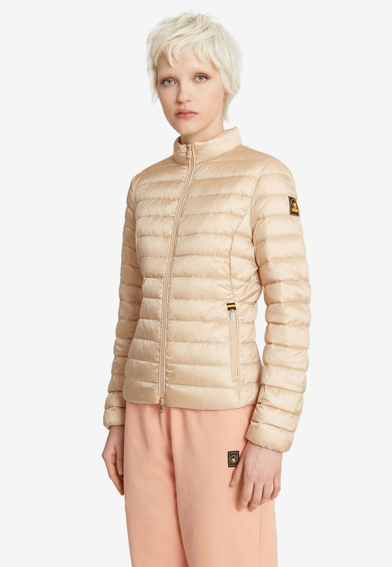 Personne aux cheveux blonds courts portant une veste matelassée beige zippée et un pantalon rose clair avec des écussons de marque visibles.