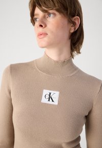 Ribovaný béžový rolák s malou bielou nášivkou loga Calvin Klein na hrudi. Mäkko textura, priliehavý dizajn.