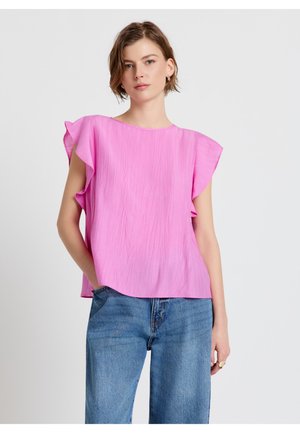 Frau trägt eine leuchtend pinke, ärmellose Bluse mit Rüschen und blaue Jeans, steht mit einer Hand in der Tasche vor einem einfarbigen Hintergrund.