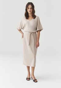 Abito beige fino al ginocchio con scollatura a V, maniche corte e cintura in vita. Tessuto liscio e design semplice. Indossato con sandali neri con tacco.