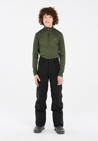 Maglione termico verde con zip abbinato a pantaloni da neve neri, caratterizzato da un design aderente e un tessuto testurizzato. Stivali grigi con suola rigata.