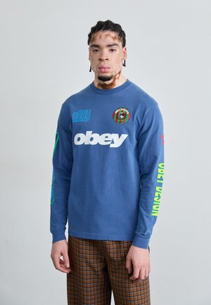 Blaues Langarm-T-Shirt mit grafischen Motiven in Weiß, Grün und Rot auf der Vorderseite und den Ärmeln. Kombiniert mit braunen karierten Hosen aus Baumwollmischung.