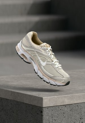 Beige en witte Nike hardloopschoen zwevend boven een betonnen richel, met zicht op het mesh bovenwerk en de details van de gevoerde zool.