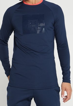 Homme portant une chemise à manches longues bleu marine avec un logo carré sombre brillant sur la poitrine et un pantalon bleu marine assorti.