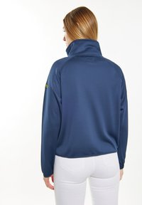 Mörkblå fleecejacka med lång ärm, hög krage och texturerat tyg; har en logotyp på vänster ärm och elastiska ärmslut.