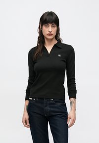 Polo nera a maniche lunghe con texture a coste, caratterizzata da un collo a camicia e un piccolo logo sul petto. Indossata con jeans denim scuro.