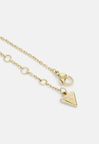 Collana in oro con maglie ovali, chiusura a moschettone e un ciondolo triangolare con la scritta "GUESS" incisa.