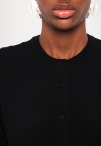 Gros plan sur le bas du visage et le cou d'une personne portant une chemise noire boutonnée, avec des lèvres visibles et un petit grain de beauté sous la bouche.