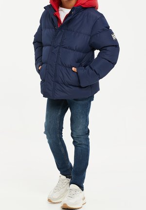 Enfant portant une doudoune bleu marine avec une doublure de capuche rouge, un jean bleu et des baskets blanches, se tenant les mains dans les poches.