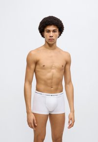 Λευκά βαμβακερά ανδρικά μποξεράκια με λωρίδα μέσης με μαύρα και κόκκινα γράμματα. Το μοντέλο στέκεται μπροστά σε ένα απλό, φωτεινό φόντο.