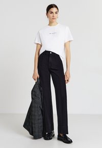 T-shirt bianca con la scritta "FRIENDS", jeans neri a vita alta, mocassini neri e una giacca grigio scuro a fantasia appoggiata su un braccio.