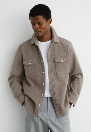 Mężczyzna w beżowej wełnianej kurtce koszulowej na guziki nałożonej na białą koszulkę i szare spodnie, ma ręce w kieszeniach, patrzy w dół stojąc na tle białej ściany.