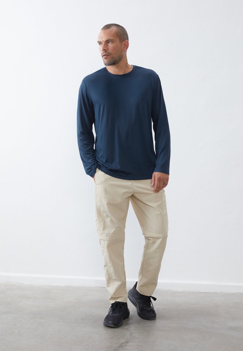 Marineblauwe longsleeve shirt, lichtbeige cargobroek met zakken, zwarte sneakers, staand tegen een witte muur, gladde textuur, casual pasvorm.