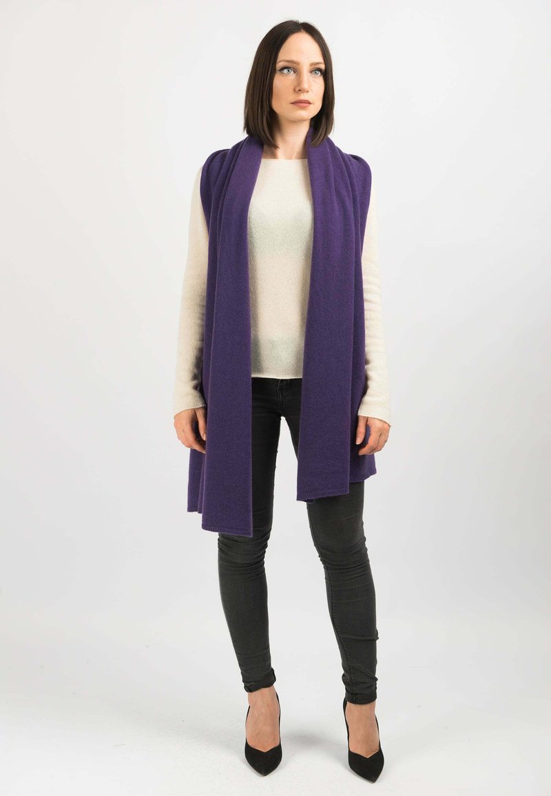 Gilet lungo viola sopra un maglione chiaro, abbinato a pantaloni skinny scuri e tacchi neri. Il tessuto appare morbido con una texture liscia.