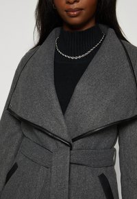 Donkere vrouw met een grijze ceintuurjas met zwarte randen over een zwart ribbeltrui en een zilveren gedraaide ketting.