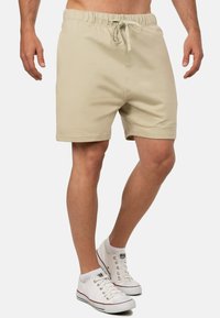 Pantaloni corti beige con vita elasticizzata e cordino; texture liscia; indossati con sneaker bianche. Nessun motivo o logo visibile.