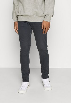 Mann trägt dunkle Slim-Fit-Jeans, weiße Sneaker mit schwarzem Nike-Logo und einen hellgrauen Sweatshirt und steht vor unifarbenem Hintergrund.