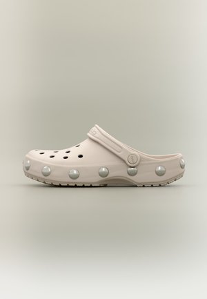 Sabot beige clair à enfiler avec des trous de ventilation et de grandes grosses clous ronds en métal autour de la semelle, avec une bride au talon.