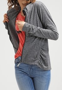 Femme portant une veste grise zippée par-dessus un t-shirt rouge à motif et un jean bleu, ajustant la fermeture éclair de la veste.