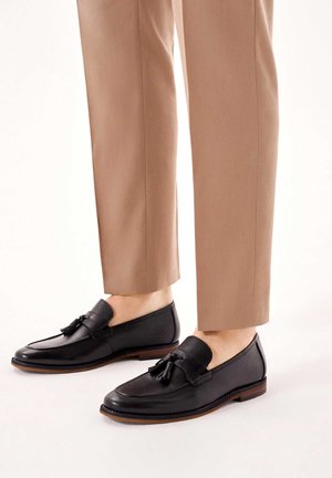 Man draagt zwarte leren tasselloafers met bruine zolen en korte lichtbruine broek op een witte achtergrond.