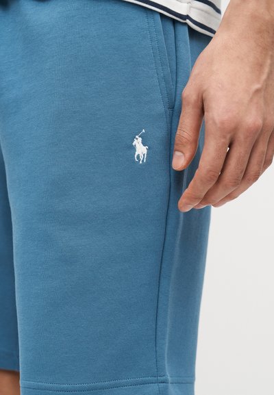 Μπλε σορτς με λευκό κεντημένο λογότυπο παίκτη πόλο στην πλαϊνή τσέπη, συνδυασμένο με χέρι που ακουμπάει δίπλα στο σορτς.