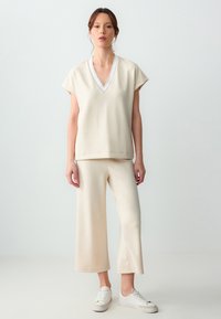 Beige V-hals top met korte mouwen, gecombineerd met wijde broek, beide van gladde stof. Witte sneakers maken de outfit compleet.