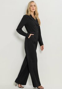 Combinaison noire ajustée avec un design croisé, des manches longues et des poches latérales. Présente des pantalons à jambes larges et une texture lisse.