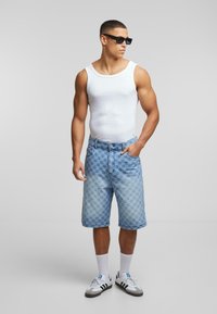 Denim shorts med et hexagonalt mønster, lys blå farve, fem lommer og en knaplukning. Båret med en hvid tanktop og solbriller.