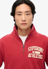 Sweatshirt zippé rouge avec un col haut, arborant un logo texturé "SUPERDRY 72 ATHLETICS" en crème sur la poitrine.