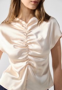 Blusa in satin a maniche corte di un delicato pesca, con dettagli arricciati sul davanti, caratterizzata da una superficie liscia e una vestibilità comoda.