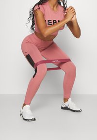 Rosa træningssæt med en crop top med sort skrift og højtaljede leggings med mesh-detaljer. Et modstandsbånd er placeret omkring benene.