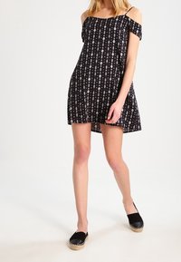 Vestido negro sin mangas con un patrón geométrico blanco, de corte corto y fabricado en tela ligera. Combinado con alpargatas negras.
