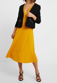 Black cropped jacket with zipper pockets, layered over a yellow dress with lace trim. Model wearing strappy black heels, standing on a plain background.