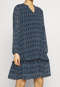 Marineblaues Kleid mit V-Ausschnitt und langen Ärmeln. Es hat ein Blattmuster in Hellblau, einen gerafften Saum und elastische Akzente an den Bündchen.