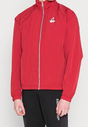 Veste rouge zippée avec col haut et petit logo noir et blanc sur la poitrine, portée par une personne portant un pantalon noir avec un texte partiel visible.