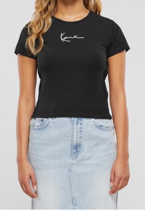 Kobieta w czarnym krótki rękaw crop top z białym kursywnym napisem i jasno niebieskiej spódnicy jeansowej z wysokim stanem.