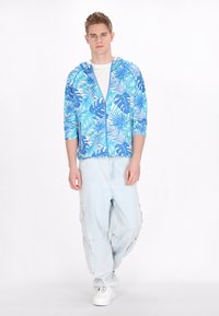 Hanorac albastru cu fermoar, cu un model tropical de frunze; material ușor. Asortat cu pantaloni cargo din denim ușori și lejeri și pantofi albi.