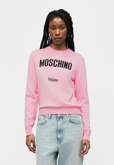 Femme aux longues tresses portant un pull Moschino Milano rose et un jean bleu clair, debout devant un fond blanc uni.