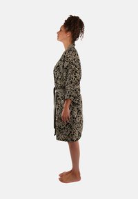 Zwarte jurk met een beige bloempatroon, voorzien van een riem en driekwart mouwen, gepresenteerd op een vrouw die blootsvoets staat, zijaanzicht.