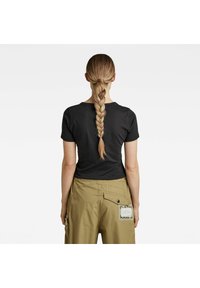 Kvinna med långt flätat hår som bär en svart t-shirt och beige byxor med bakficka och applikation, som står framför en enkel vit bakgrund.