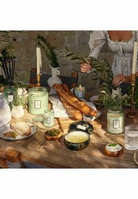 Träbord uppsatt med gröna ljus, färskt bröd, burkar och tallrikar; har texturerade material, jordnära färger och dekorativa detaljer.