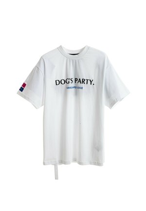 Joven mujer de pie con una camiseta blanca de gran tamaño con el texto "DOG'S PARTY", pantalones cortos blancos, zapatos slip-on texturizados y sosteniendo una etiqueta negra.