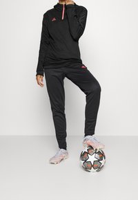 Haut de survêtement noir à zip avec des accents roses, associé à un pantalon noir fuselé et des crampons de football gris clair. Un ballon de football est posé au sol.