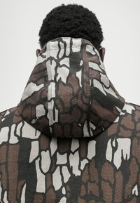 Kapuzensweatshirt mit einem strukturierten, abstrakten Muster in Braun, Weiß und Schwarz. Sichtbare Nähte und ein lässiges Kapuzen-Design.