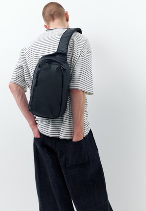 Uomo con borsa a tracolla nera, camicia a righe bianche e nere e pantaloni scuri, in piedi con le mani nelle tasche posteriori.