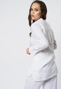 Camicia bianca in lino con maniche lunghe, colletto a punta e fessure laterali. Il tessuto presenta una leggera texture, con una vestibilità comoda e dettagli di piega sulla parte posteriore.