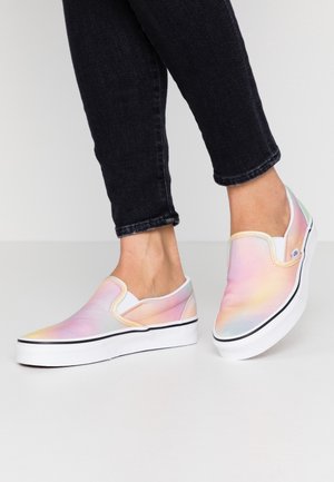 Osoba nosząca pastelowe tęczowe trampki typu slip-on z białymi podeszwami i czarną lamówką, w połączeniu z czarnymi, przylegającymi jeansami z mankietami.
