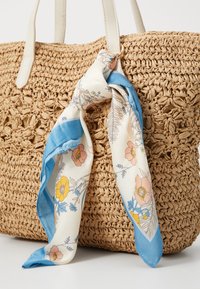 Sac cabas en paille tressée, fabriqué en matériau beige, avec des poignées blanches robustes et un foulard en soie décoratif à motifs floraux en bleu, rose et jaune.